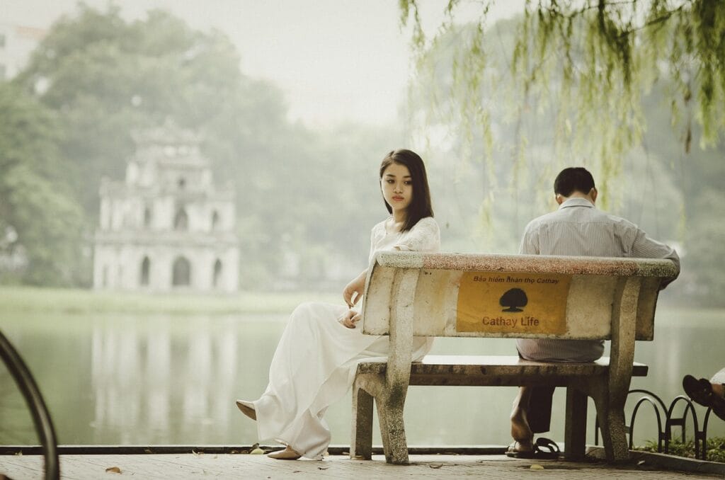 Ek udaas ladki aur uska door ho chuka saathi jheel ke kinare ek bench par baithe hain, jahan samay ke saath badalte relationship ka dard aur ehsaas chhupa hai.